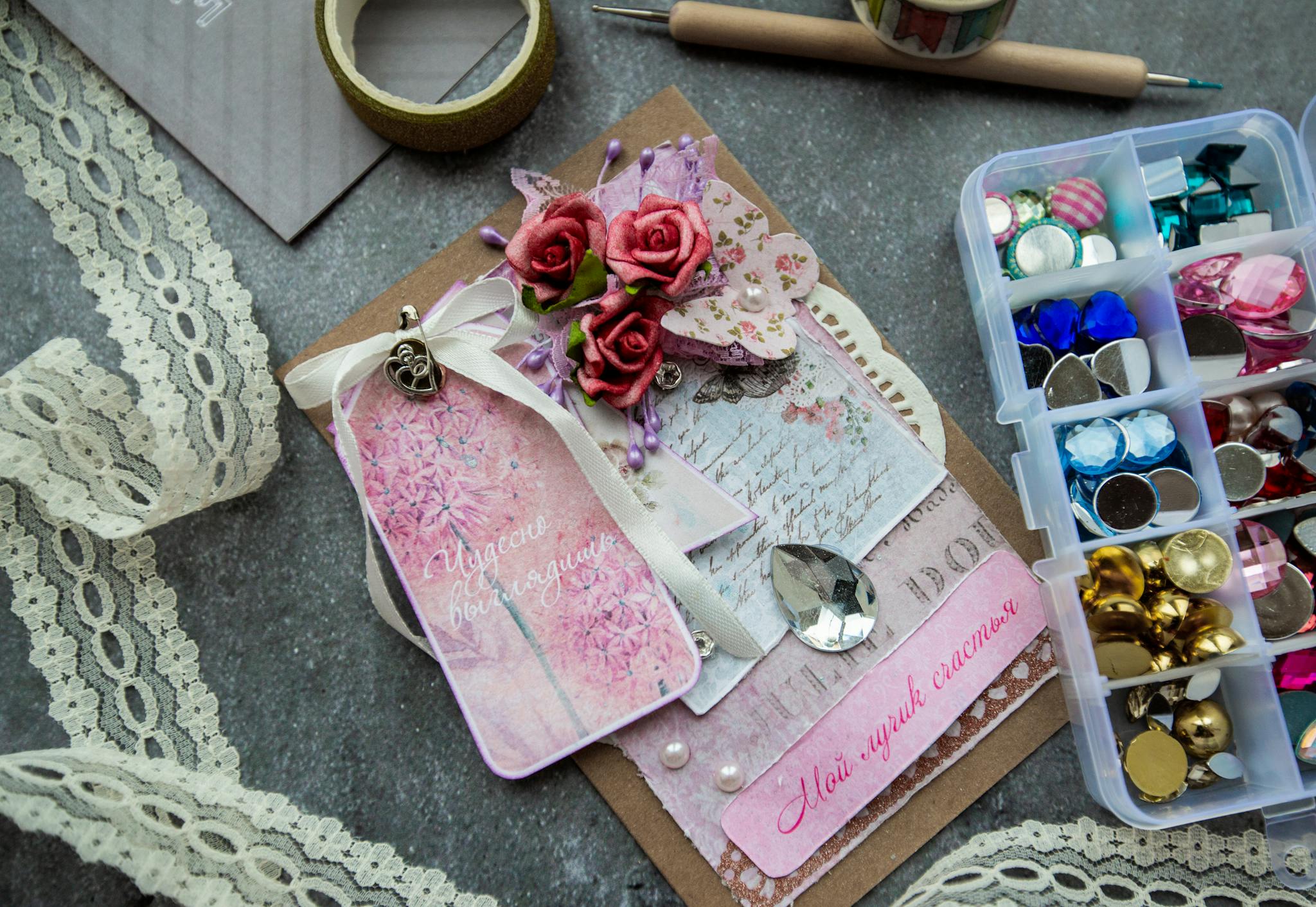 Top view of handicraft greeting card decorated with artificial flowers and fancy stones placed on table near box of colorful beads and buttons and tape lace